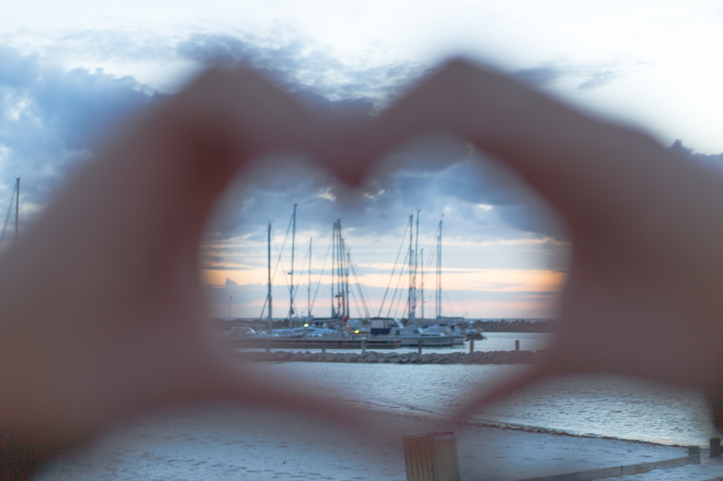 Two hands creating the shape of a heart with a harbor and a sunset in the background.