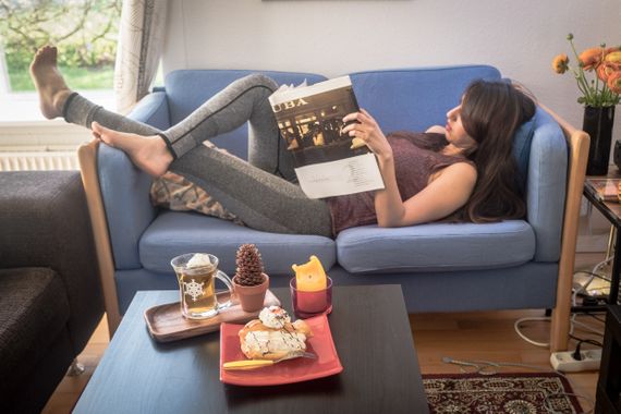 Arina lying on a blue couch reading a magazine with a cake and a cup of tea on the table in front of her