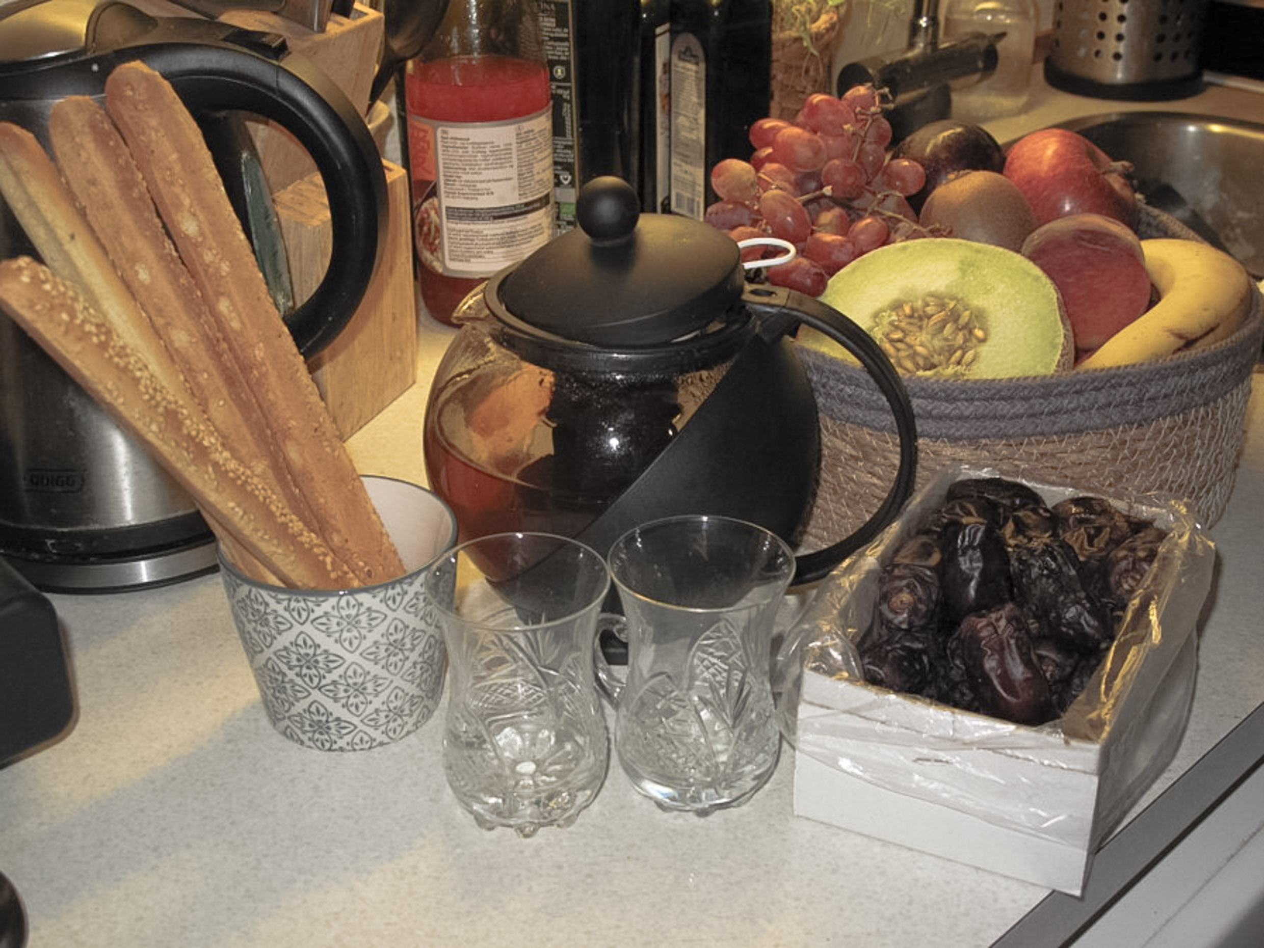 A table with a bowl of fruit, two glasses, a tea pot, bread sticks and dates.