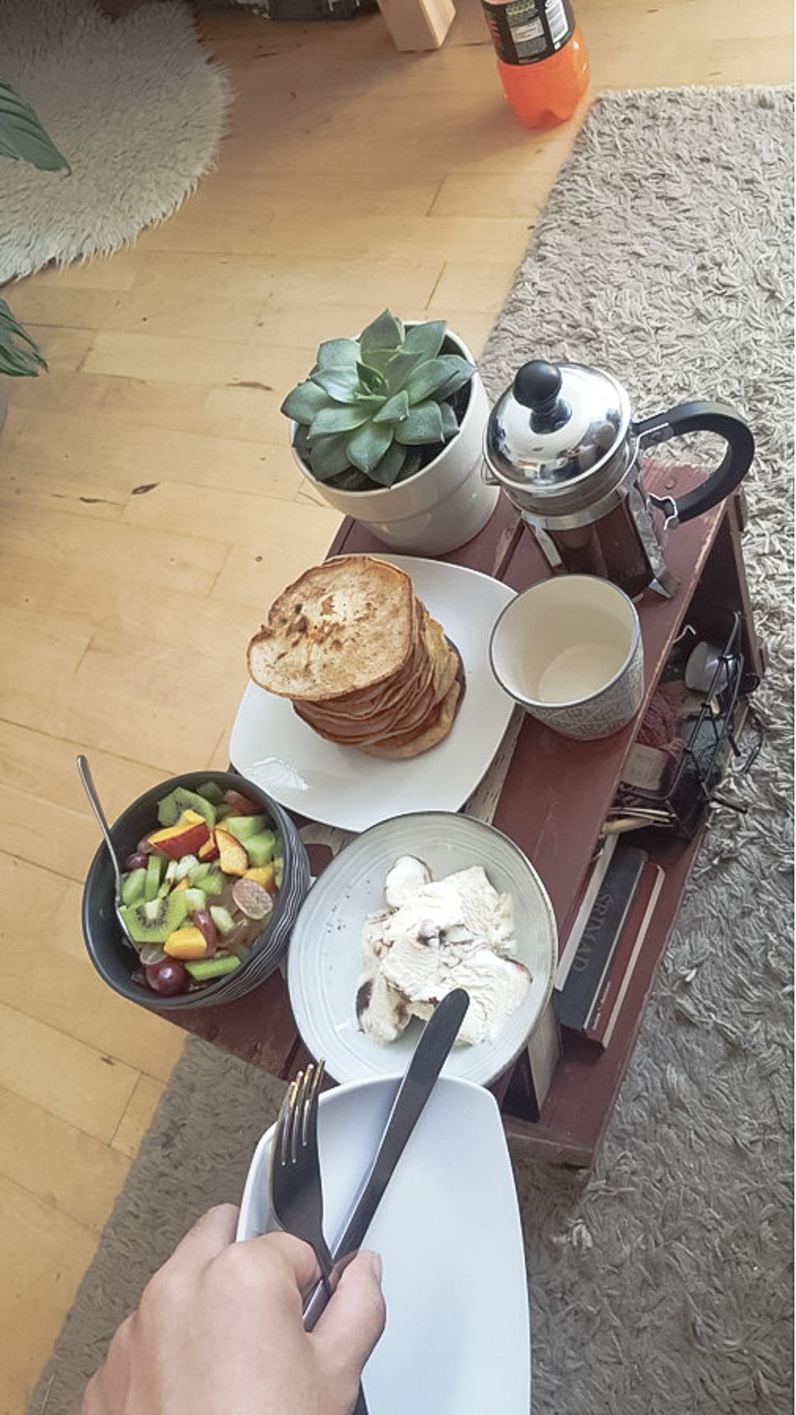 A table with a plant, coffee, pancakes, a bowl of fruit and icecream.
