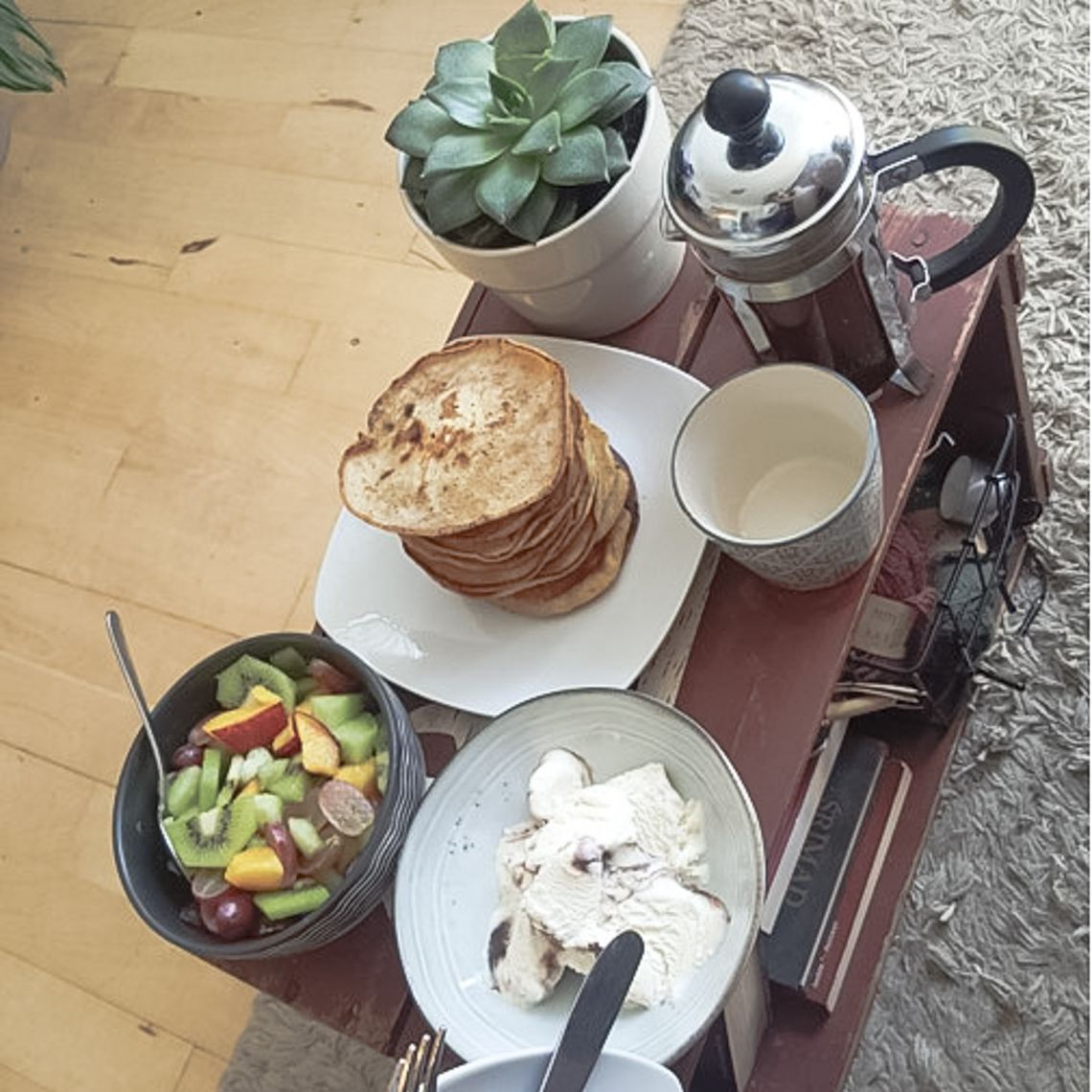 A table with a plant, coffee, pancakes, a bowl of fruit and icecream.