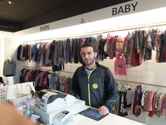 Abdulsalam standing behind a cash register with rows of baby and childrens clothes hanging in the background