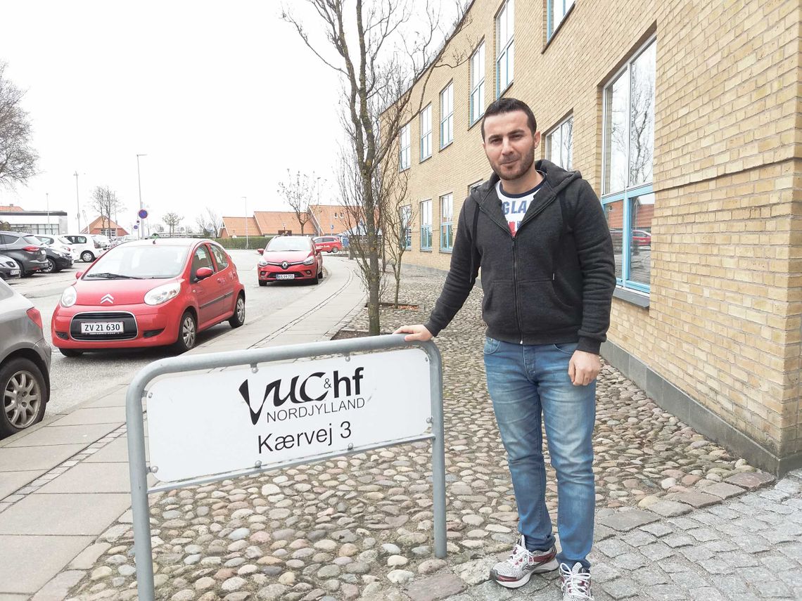 Abdulsalam leaning against a sign saying VUC & hf Nordjylland, Kærvej 3, in front of a yellow brick building and parked cars