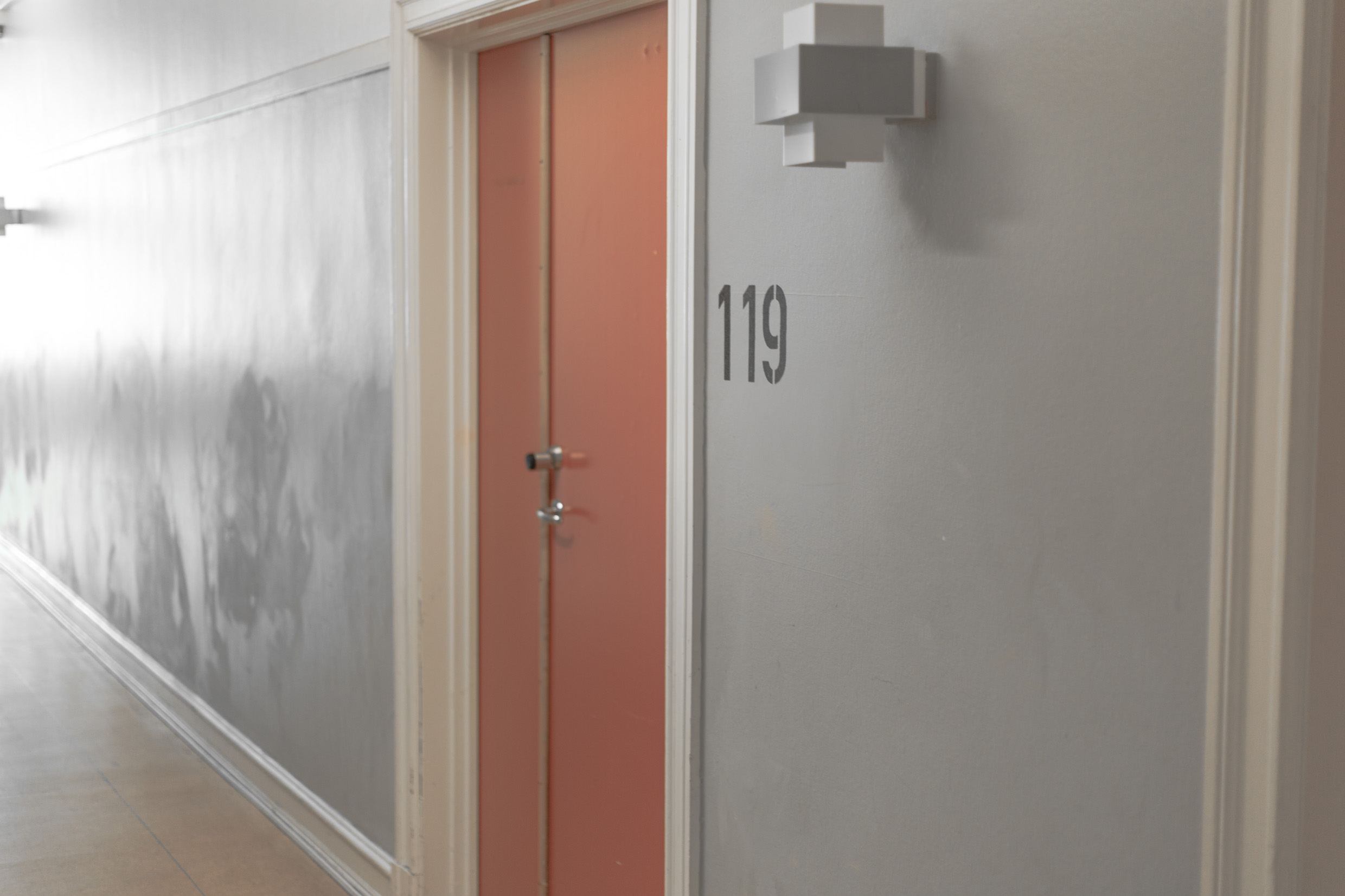 A red door in a grey hallway with the number 119 written in black letters next to it.