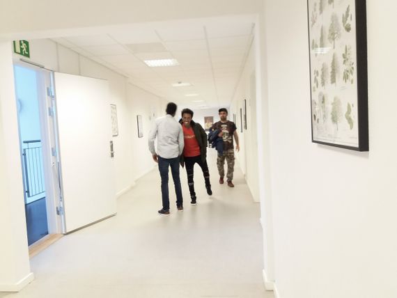 Two guys greeting each other with a handshake on a white hallway, while another man is walking in the background