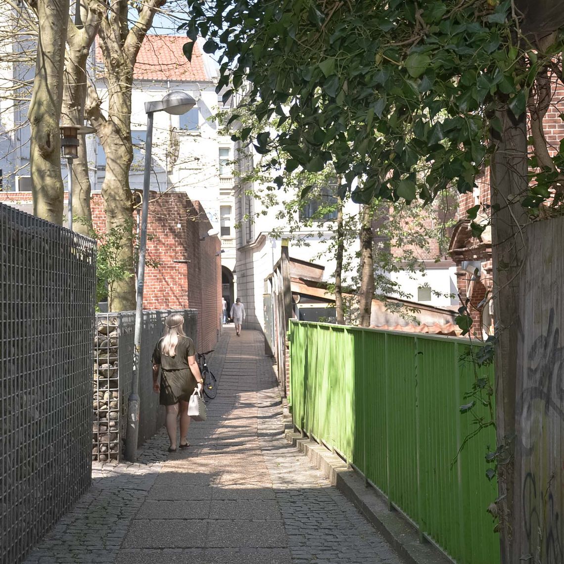 An alley with a green fence and a woman walking away from the camera