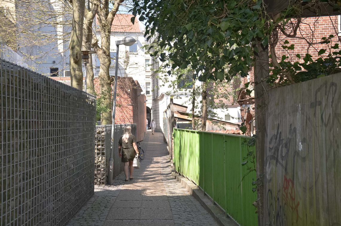 An alley with a green fence and a woman walking away from the camera