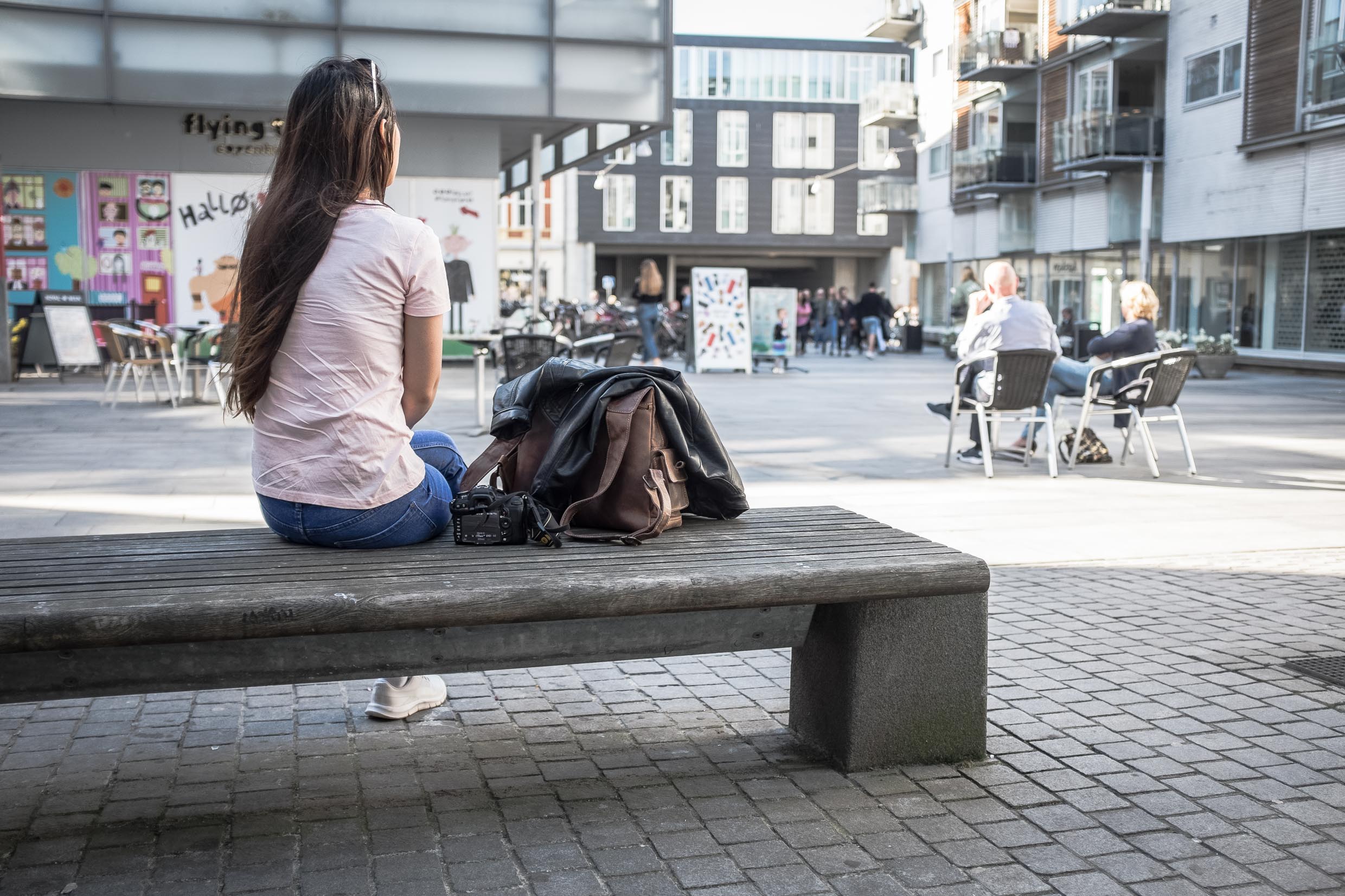 Arina is sitting on a bench with her bag next to her, looking at people walking and sitting in the street