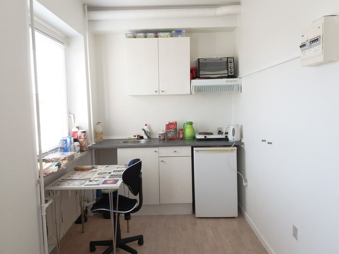 A kitchen with a small table, a blue chair, a zink, microwave, a boiler and a fridge.