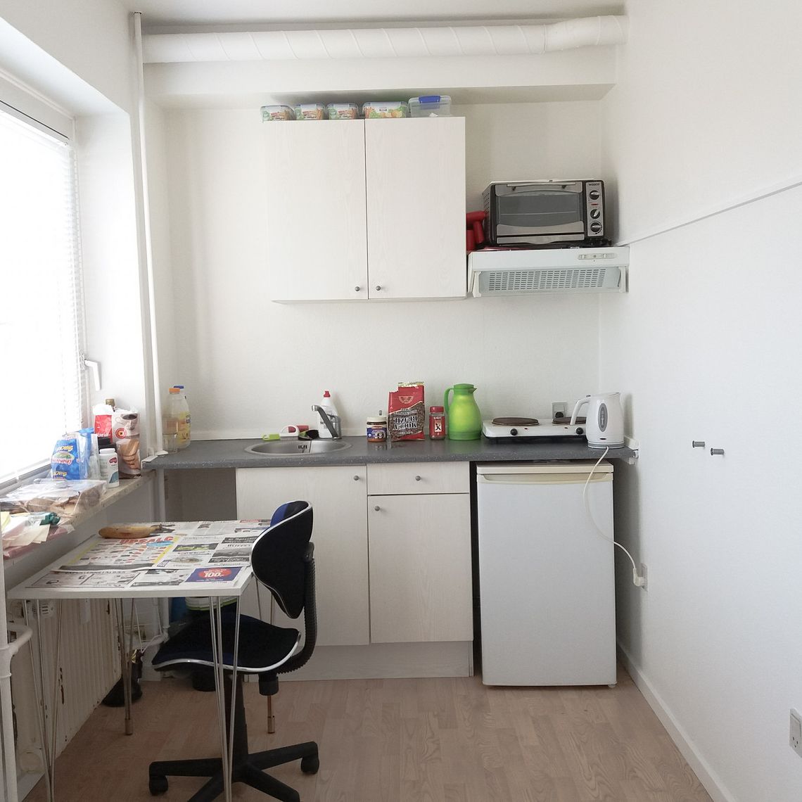 A kitchen with a small table, a blue chair, a zink, microwave, a boiler and a fridge.