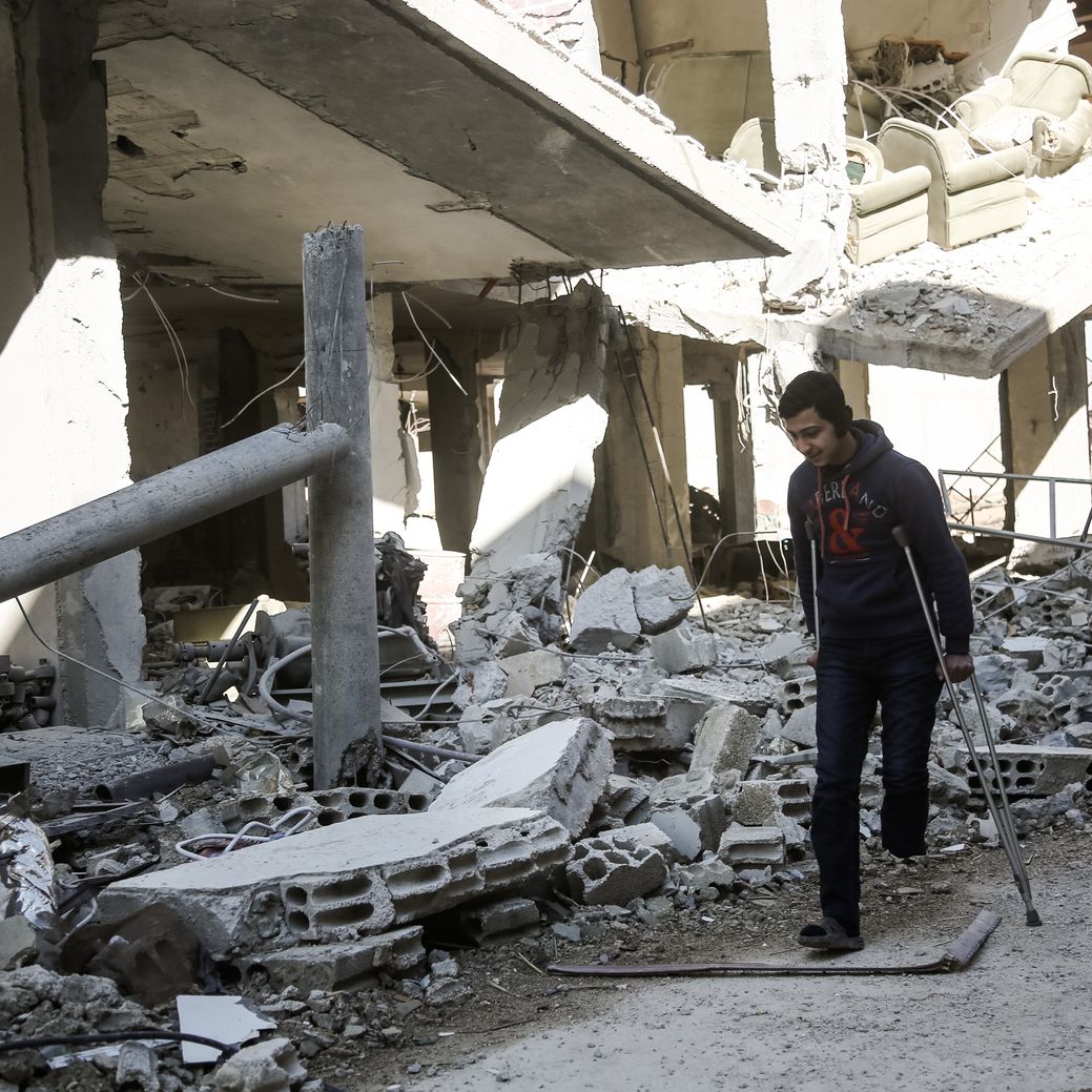 Photo by Salem Mdlala. A young man walking with crutches next to a destroyed building in Ghouta.