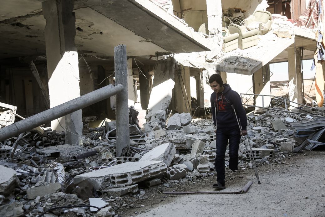Photo by Salem Mdlala. A young man walking with crutches next to a destroyed building in Ghouta.