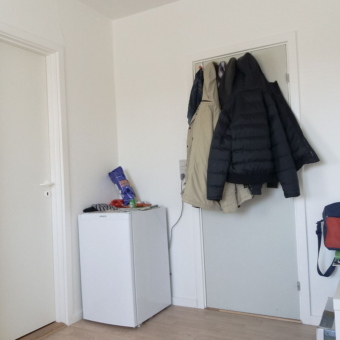 A door with a few jackets haning on it, next to a chair with a red and blue bag on top.