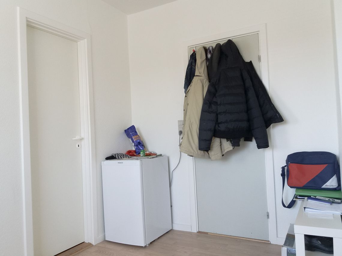 A door with a few jackets haning on it, next to a chair with a red and blue bag on top.