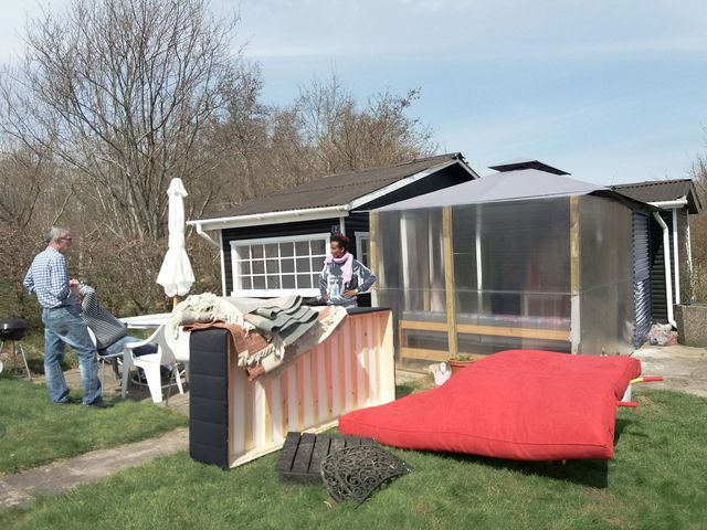 A big red mattress, a bed and three people standing on the grass outside a small black house with white windows