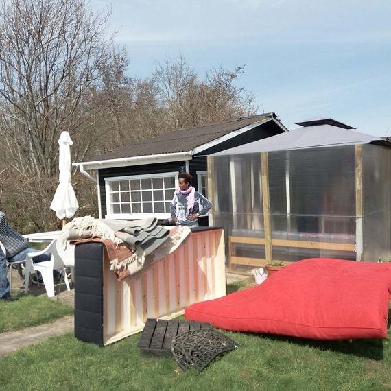 A big red mattress, a bed and three people standing on the grass outside a small black house with white windows