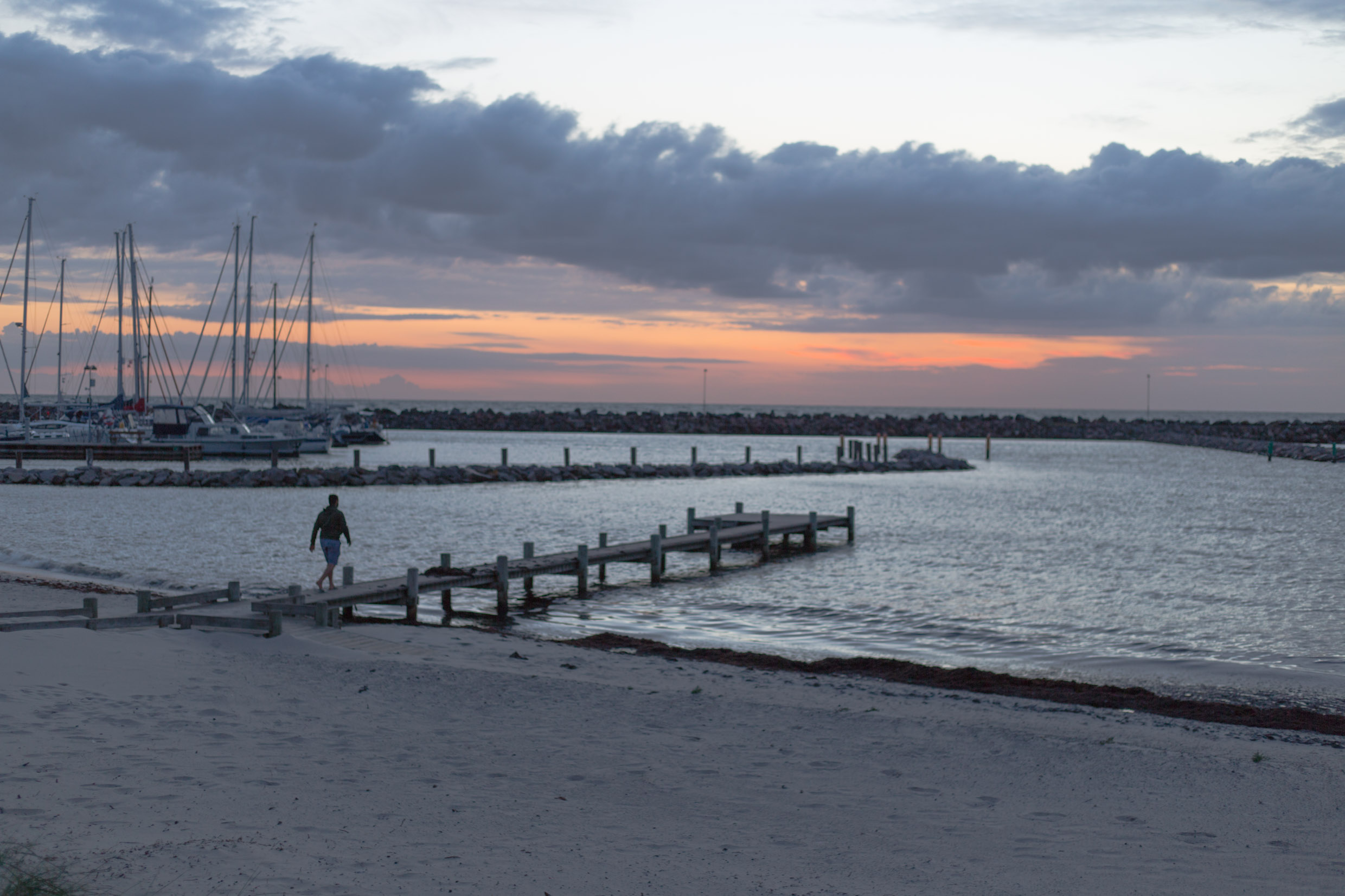 Hisham walking on a jetty on a beach near a harbor in the sunset.