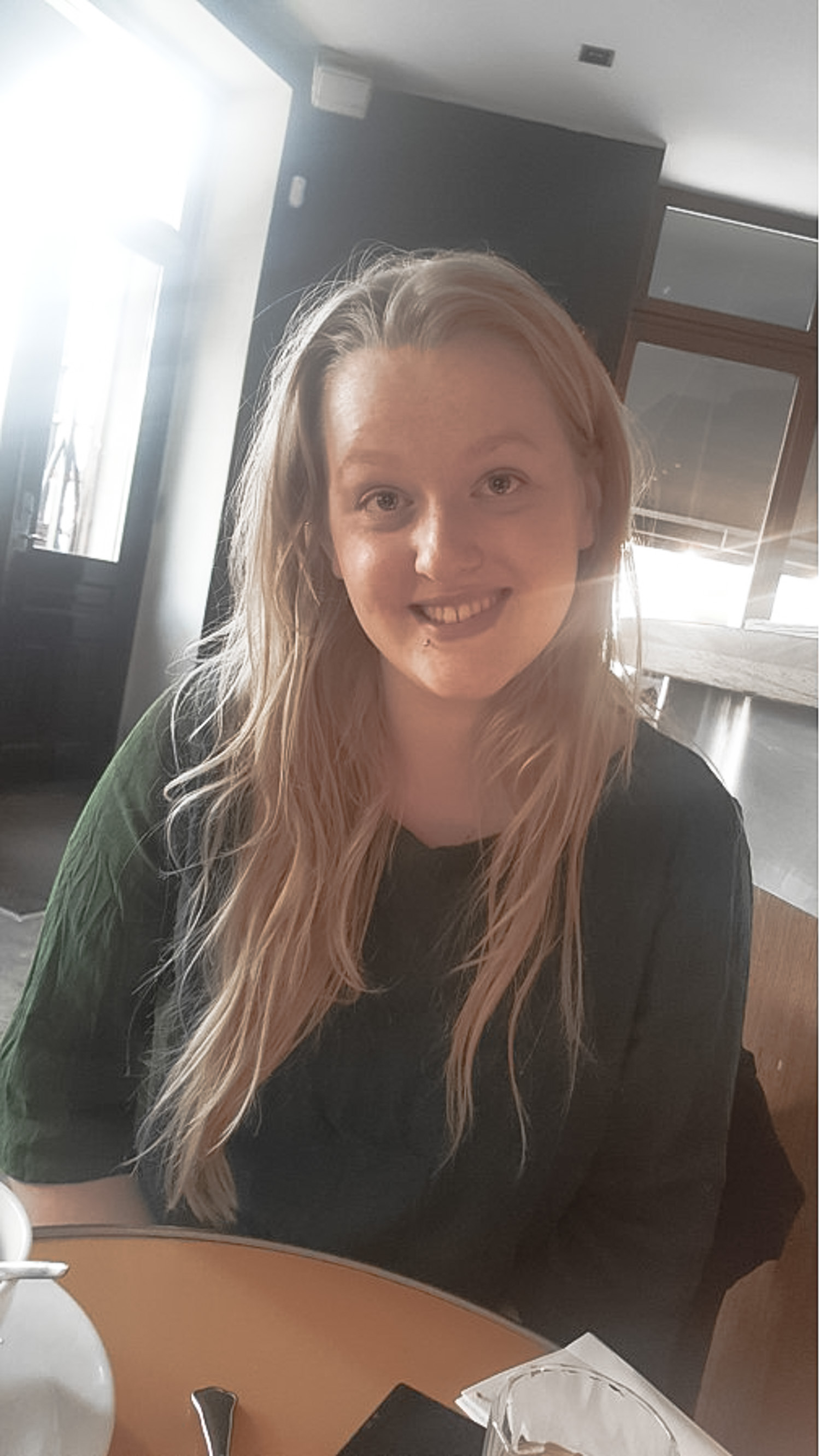 A girl with blond hair and a green sweater, sitting at a table and smiling to the camera.