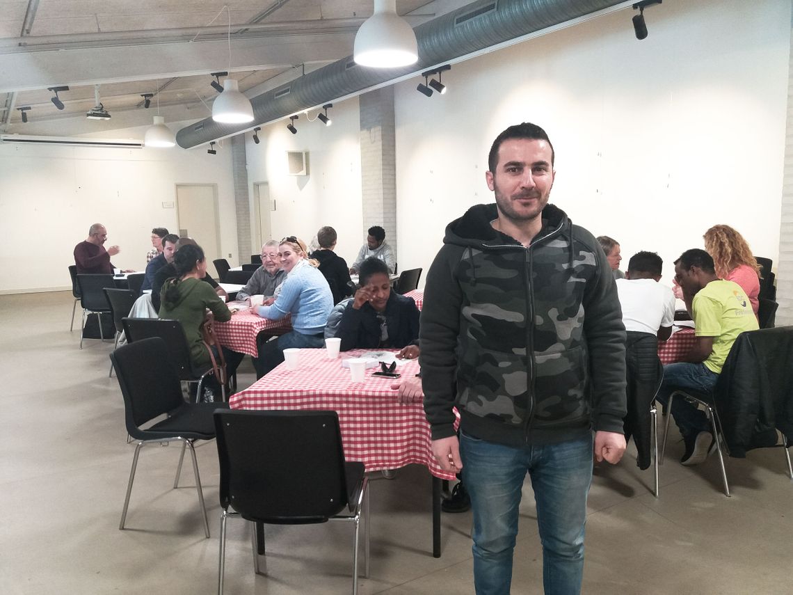 Abdulsalam standing in a big room, with tables with a red and white checkered tablecloth, while people are sitting around the tables talking and doing homework