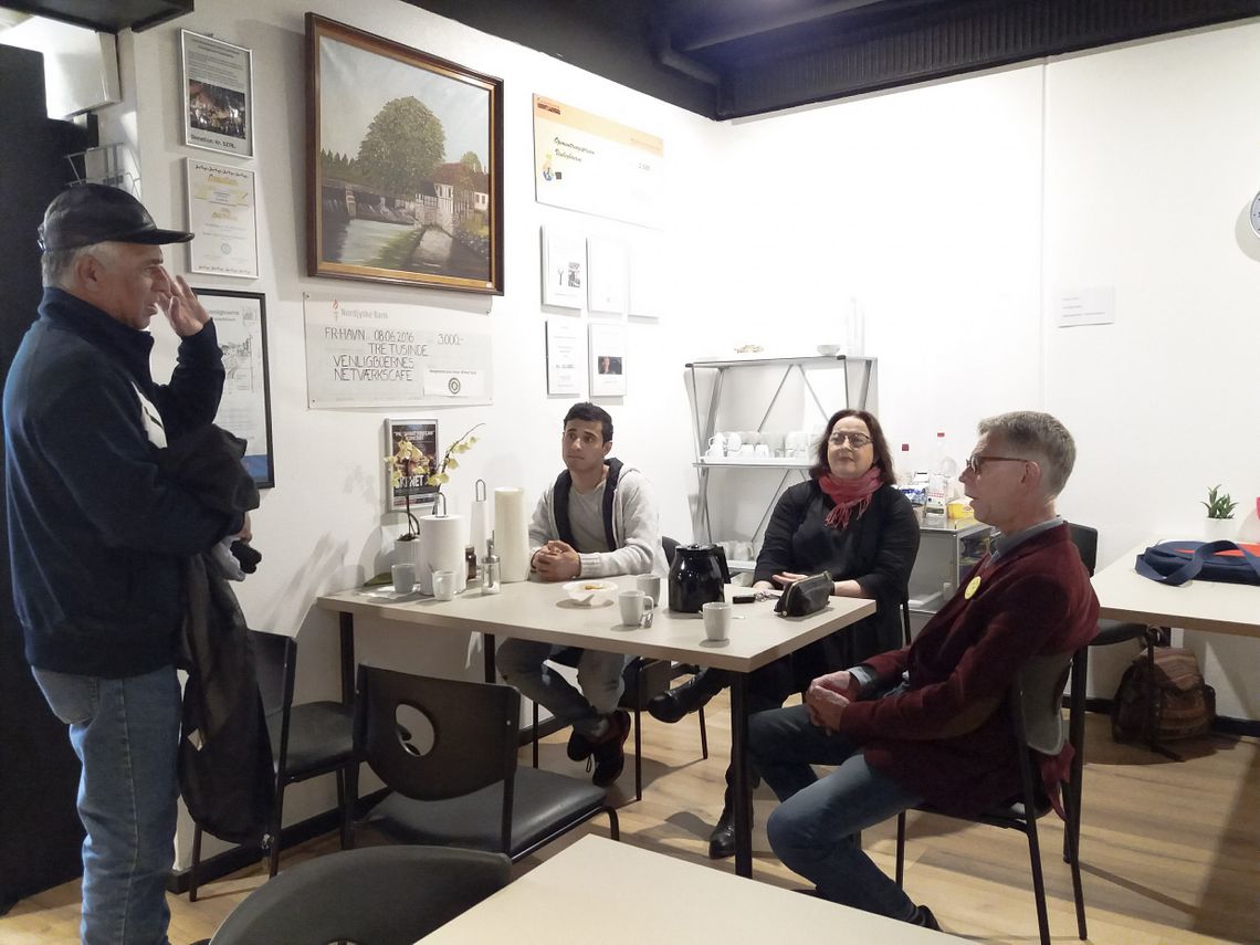 A man standing, holding his jacket, while wawing to three people sitting around a white table with cups on it