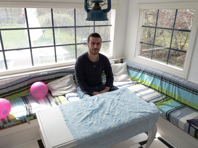 Abdulsalam sitting on a bench with colored cushions, two baloons and two big windows in the background