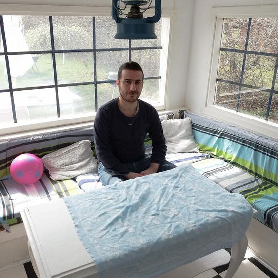 Abdulsalam sitting on a bench with colored cushions, two baloons and two big windows in the background