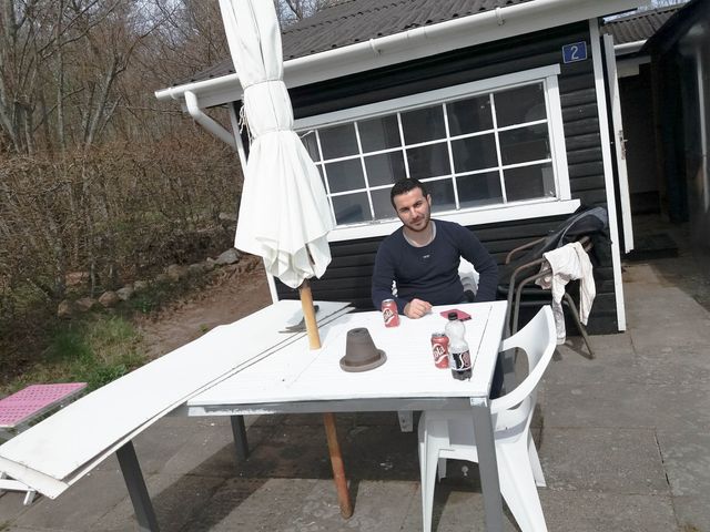 Abdulsalam sitting at a white table, with a parasol, outside a small black house with white windows