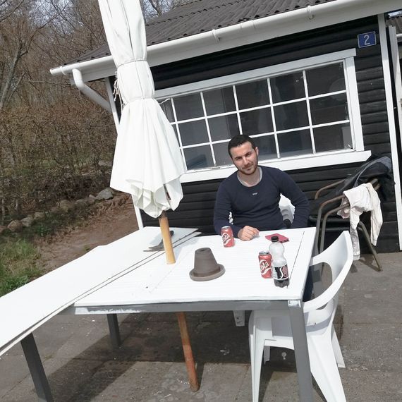 Abdulsalam sitting at a white table, with a parasol, outside a small black house with white windows