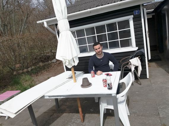 Abdulsalam sitting at a white table, with a parasol, outside a small black house with white windows