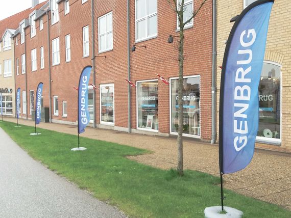 Four blue flags saying "Genbrug" standing on the grass outside of a big red brick building